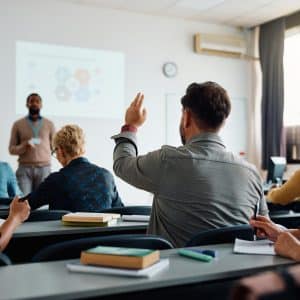 Back view of older student raising his hand to answer teacher's question during education training class.