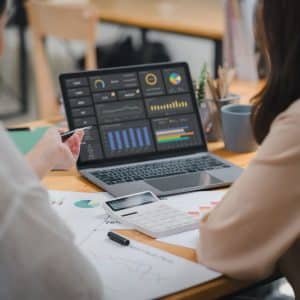 Two businesspeople or an accountant team are analyzing data charts, graphs, and a dashboard on a laptop screen in order to prepare a statistical report and discuss financial data in an office.